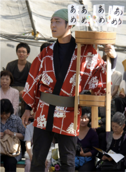 A performer golds a wooden stool marked with kanji flags. Street vendors and performers depicted in the speculative historical theater, written by Hayashi.