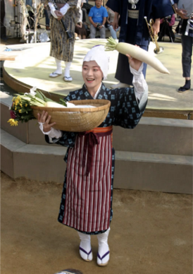 A performer holds a giant vegetable above her head with one hand, and holds a large wicker basket in her other arm. Street vendors and performers depicted in the speculative historical theater, written by Hayashi.