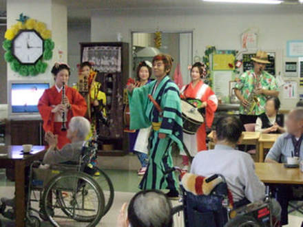 Chindon Tsushinsha visiting an old people's home. Perfomers play instruments dressed in colorful robes. Older adults sit at tables listening, some in wheelchairs.