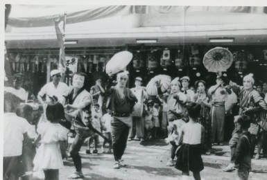 Black and white photograph; chindon-ya troupe holding parasols while children watch the performance.